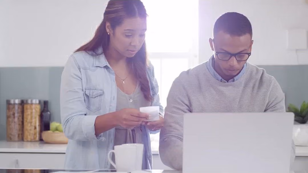 Young couple in their kitchen looking over a laptop together, looking concerned.