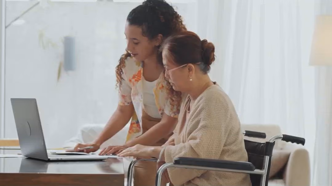 Young woman helping an older woman in a wheelchair with something on a laptop.
