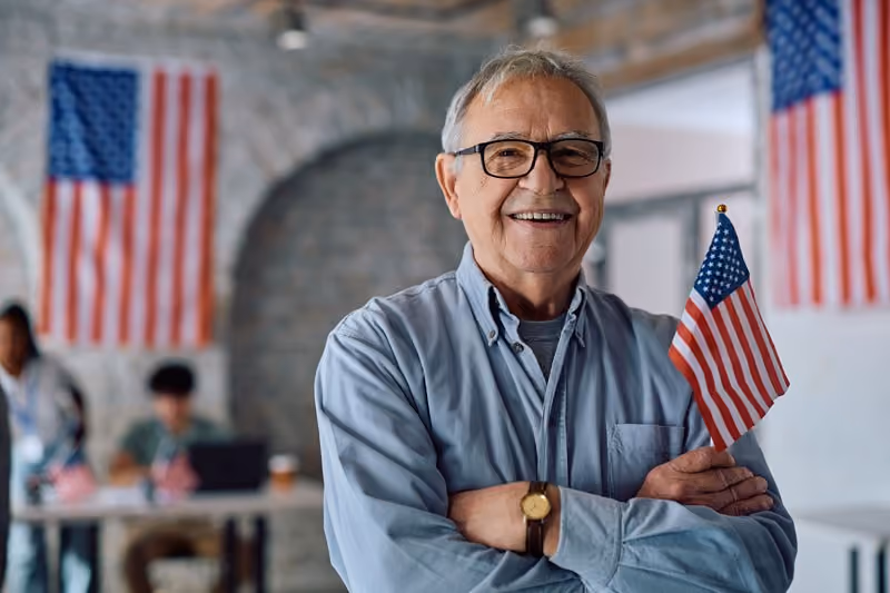 Smiling older man holding a small American flag, with flags on the wall behind him.